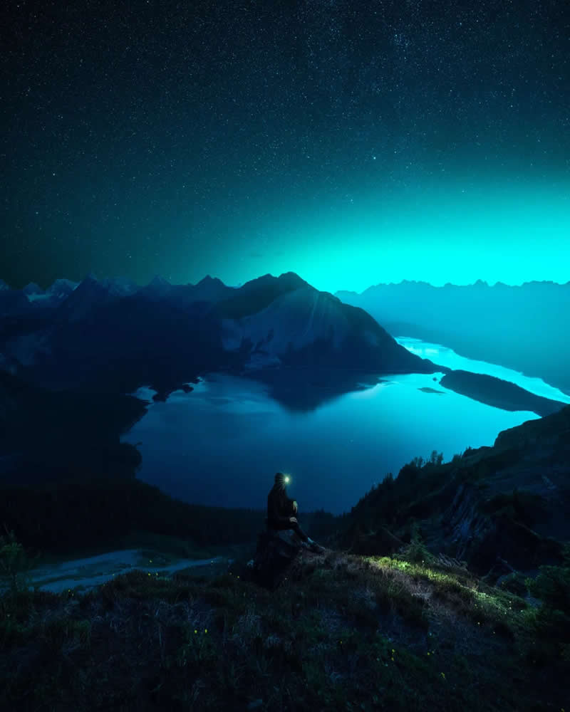 Blue Nights and Snowy Peaks: Cath Simard's 25 Dreamlike Landscapes of the Canadian Rockies 45 A lone hiker with a headlamp overlooks glowing alpine lakes and dark mountain silhouettes beneath a star-filled night sky, creating a peaceful and atmospheric Canadian Rockies landscape.