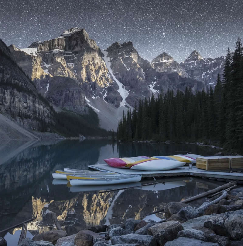 Blue Nights and Snowy Peaks: Cath Simard's 25 Dreamlike Landscapes of the Canadian Rockies 46 Colorful canoes rest on a wooden dock beside a calm alpine lake, reflecting towering snow-capped mountains, evergreen forests, and a star-filled night sky in the Canadian Rockies.