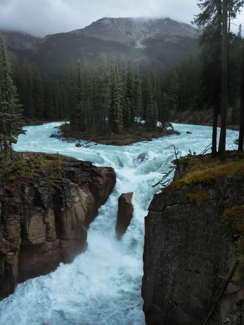 Blue Nights and Snowy Peaks: Cath Simard's 25 Dreamlike Landscapes of the Canadian Rockies 47 A powerful glacial river crashes through a rocky canyon below a forested island, surrounded by evergreen trees and misty mountains in the Canadian Rockies under an overcast sky.