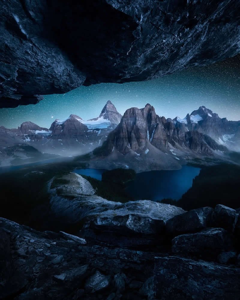 Blue Nights and Snowy Peaks: Cath Simard's 25 Dreamlike Landscapes of the Canadian Rockies 30 A star-filled night sky illuminates the Canadian Rockies, viewed through the opening of a rocky cave overlooking a deep blue alpine lake and dramatic snow-capped mountain peaks.