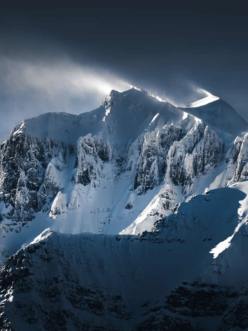 Blue Nights and Snowy Peaks: Cath Simard's 25 Dreamlike Landscapes of the Canadian Rockies 48 A close-up view of snow-covered mountain peaks with windblown spindrift sweeping across the summits, highlighting rugged cliffs, icy textures, and dramatic light in the Canadian Rockies.