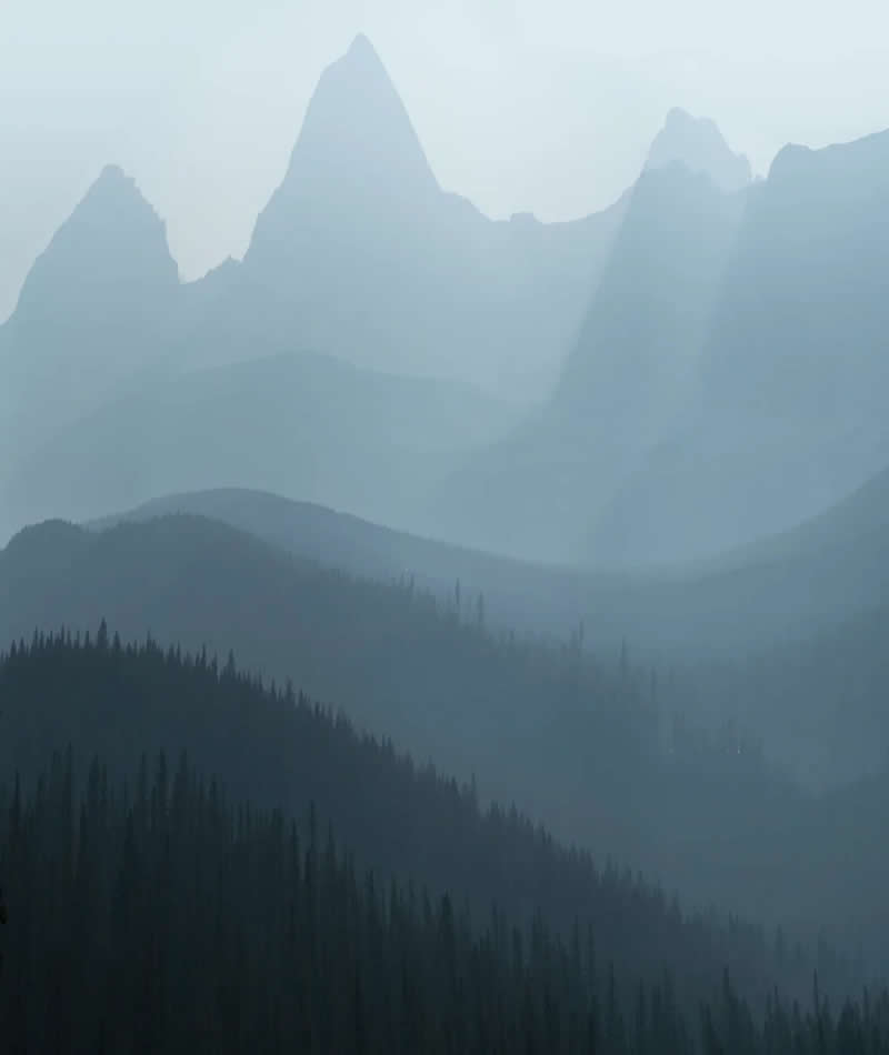 Blue Nights and Snowy Peaks: Cath Simard's 25 Dreamlike Landscapes of the Canadian Rockies 51 Misty mountain peaks rise above layered forested ridges in the Canadian Rockies, with soft atmospheric haze creating depth, silhouettes, and a peaceful minimalist landscape.