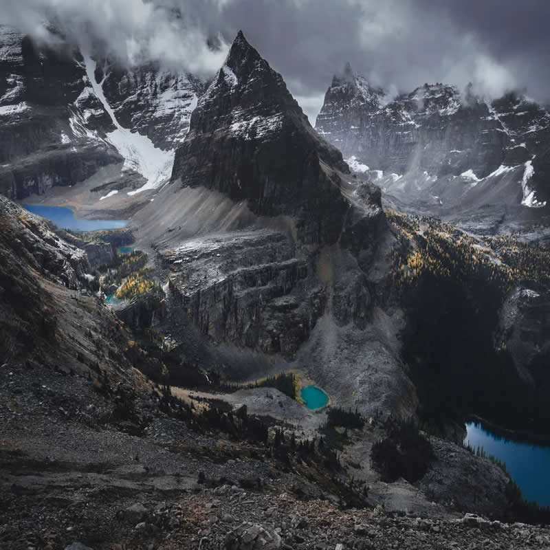 Blue Nights and Snowy Peaks: Cath Simard's 25 Dreamlike Landscapes of the Canadian Rockies 52 Towering jagged mountain peaks rise above turquoise glacial lakes and rocky valleys, with low clouds swirling around snow-covered summits in the dramatic Canadian Rockies landscape.
