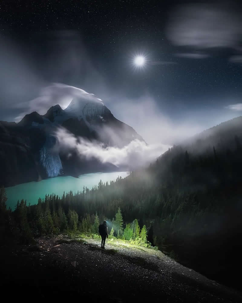 Blue Nights and Snowy Peaks: Cath Simard's 25 Dreamlike Landscapes of the Canadian Rockies 31 A lone hiker overlooks a turquoise glacial lake beneath moonlit, mist-covered mountains in the Canadian Rockies, with evergreen forests and a star-filled night sky creating a dramatic alpine landscape.