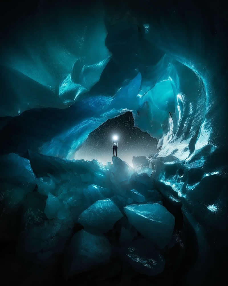Blue Nights and Snowy Peaks: Cath Simard's 25 Dreamlike Landscapes of the Canadian Rockies 32 A solitary explorer with a bright headlamp stands inside a glowing blue ice cave, surrounded by massive sculpted ice formations and crystalline textures in a dramatic frozen landscape.
