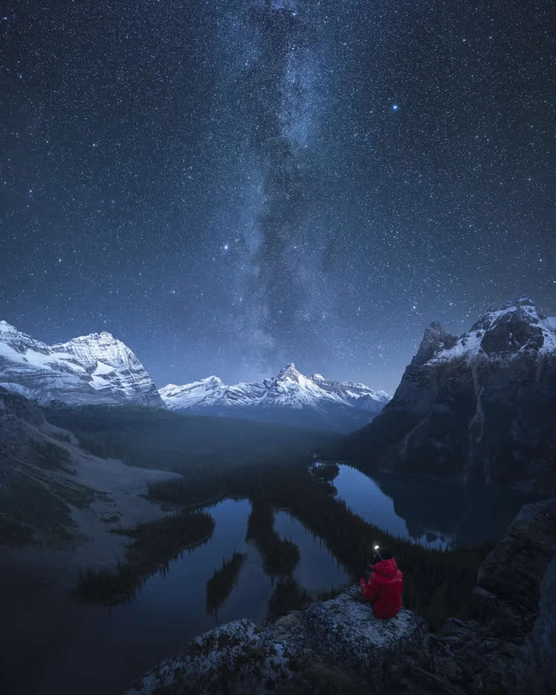 Blue Nights and Snowy Peaks: Cath Simard's 25 Dreamlike Landscapes of the Canadian Rockies 33 A lone person in a red jacket sits on a rocky overlook beneath the Milky Way, gazing across winding lakes and snow-capped peaks in the Canadian Rockies at night.