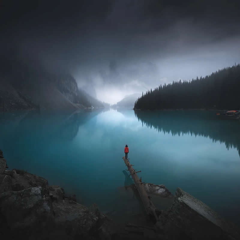 Blue Nights and Snowy Peaks: Cath Simard's 25 Dreamlike Landscapes of the Canadian Rockies 35 A lone person in a red jacket stands on a fallen log extending into a turquoise glacial lake, surrounded by misty mountains, evergreen forests, and calm reflections in the Canadian Rockies.