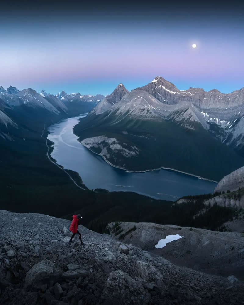 Blue Nights and Snowy Peaks: Cath Simard's 25 Dreamlike Landscapes of the Canadian Rockies 36 A lone hiker in a red jacket ascends a rocky slope overlooking a winding alpine lake, surrounded by towering Canadian Rockies beneath a pastel twilight sky and a glowing moon.