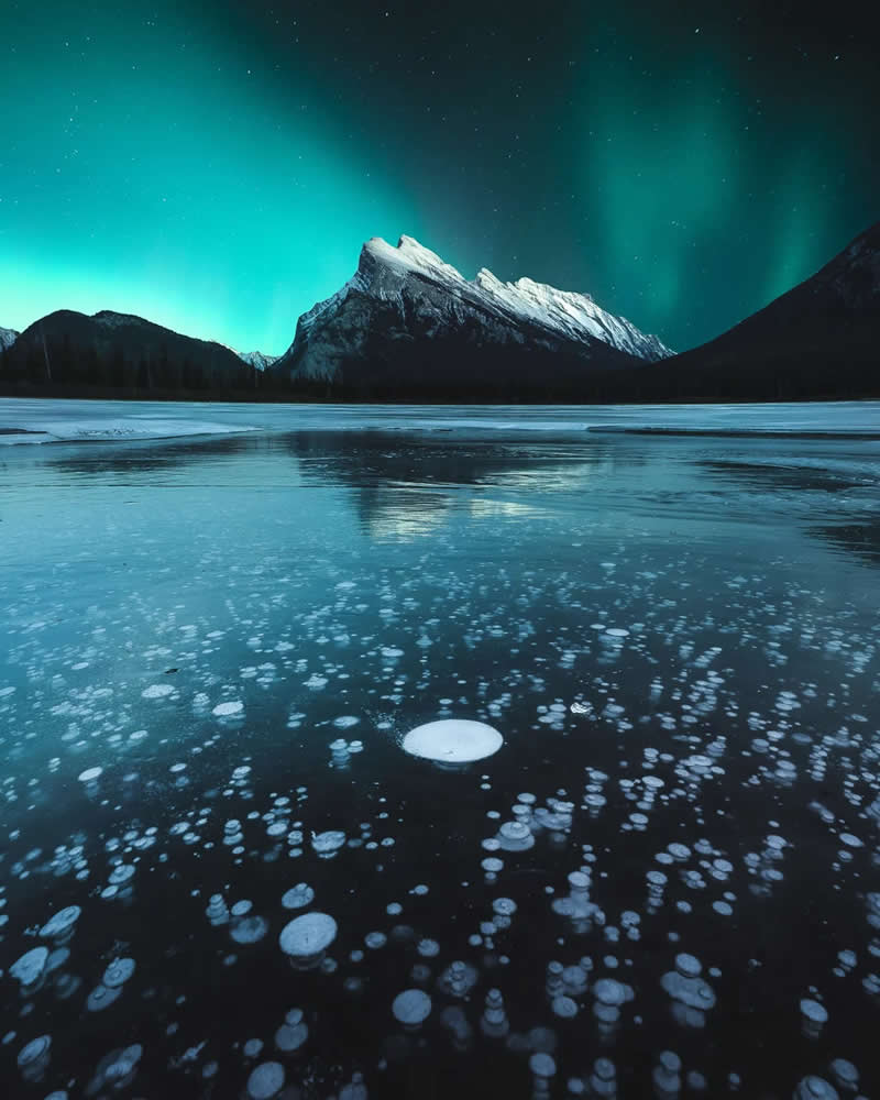 Blue Nights and Snowy Peaks: Cath Simard's 25 Dreamlike Landscapes of the Canadian Rockies 37 Green aurora borealis glows above a snow-covered mountain reflected on a frozen lake filled with trapped ice bubbles, creating a striking winter landscape in the Canadian Rockies.