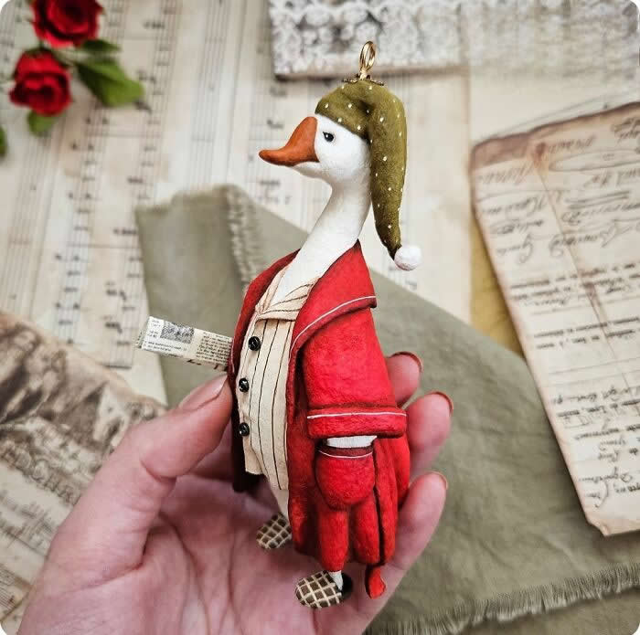 Miniature World: 33 Cotton Wool Sculptures by Katya Celebrating the Magic of Handmade Craft 39 Cotton Wool Sculptures. A handcrafted miniature goose ornament made from cotton wool is dressed in a deep red coat and striped vest, wearing a soft green nightcap while holding a tiny rolled paper scroll against a vintage-style holiday background.