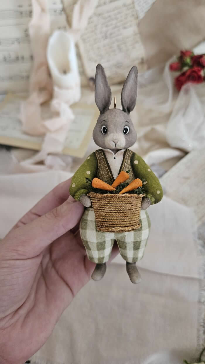 Miniature World: 33 Cotton Wool Sculptures by Katya Celebrating the Magic of Handmade Craft 48 A handcrafted miniature rabbit ornament made from cotton wool wears a soft green jacket and checkered trousers while holding a woven basket filled with tiny carrots, posed against a delicate vintage-inspired background.