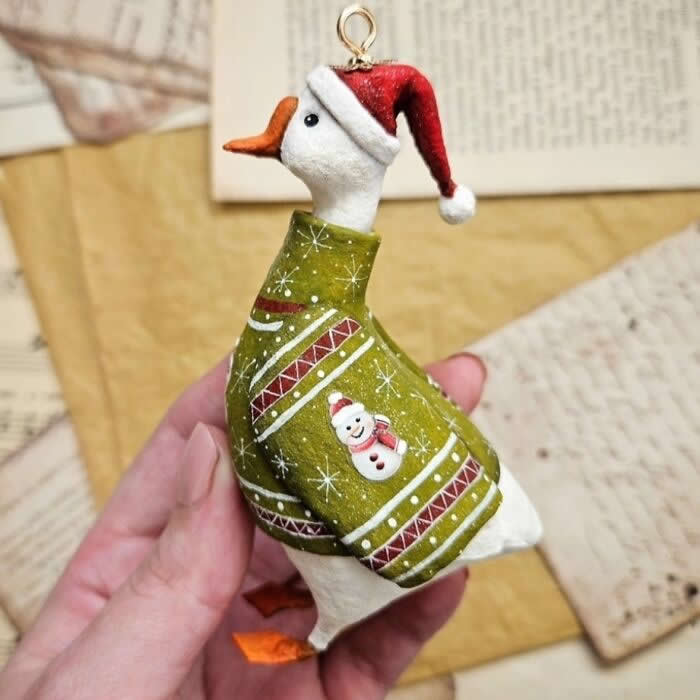 Miniature World: 33 Cotton Wool Sculptures by Katya Celebrating the Magic of Handmade Craft 50 A handcrafted miniature goose ornament made from cotton wool wears a cozy olive-green Christmas sweater with festive patterns and a tiny snowman design, paired with a red Santa hat, posed against a vintage paper background.