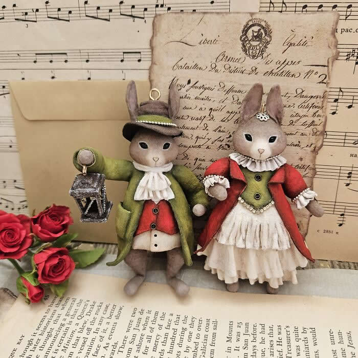 Miniature World: 33 Cotton Wool Sculptures by Katya Celebrating the Magic of Handmade Craft 51 Two handcrafted miniature rabbit ornaments made from cotton wool stand together against a vintage backdrop of sheet music, old letters, and red roses—one dressed in a green coat holding a lantern, the other in a red coat and layered cream dress.