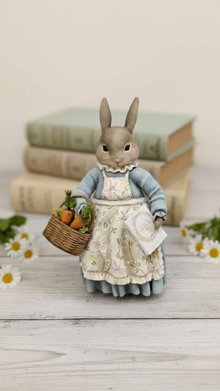 Miniature World: 33 Cotton Wool Sculptures by Katya Celebrating the Magic of Handmade Craft 55 A handcrafted miniature rabbit ornament made from cotton wool wears a soft blue dress and floral apron, carrying a woven basket of tiny carrots and a small letter, posed in front of stacked vintage books and scattered daisies.