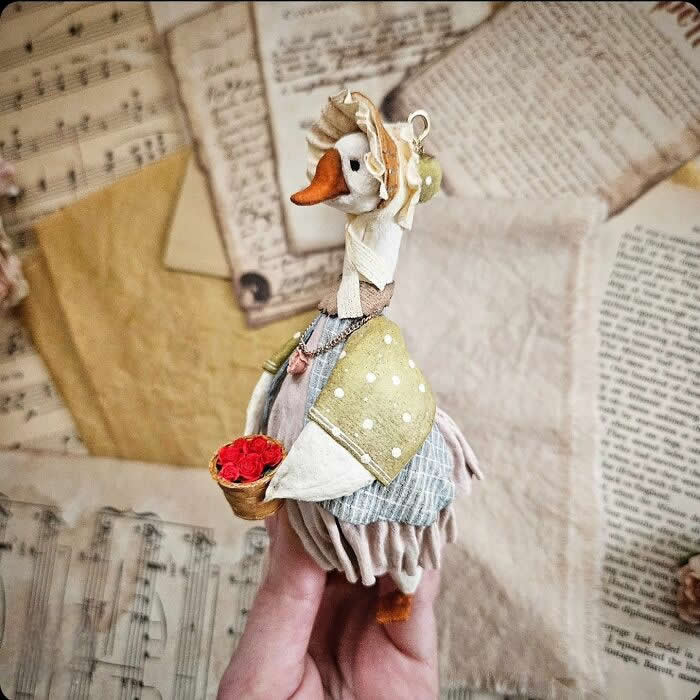 Miniature World: 33 Cotton Wool Sculptures by Katya Celebrating the Magic of Handmade Craft 56 A handcrafted miniature goose ornament made from cotton wool wears a soft bonnet, polka-dot shawl, and layered vintage dress while carrying a tiny basket of red roses, posed against a backdrop of old sheet music and aged paper.