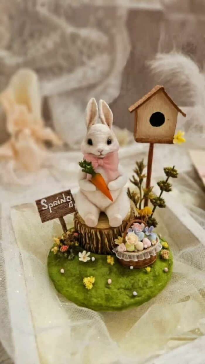 Miniature World: 33 Cotton Wool Sculptures by Katya Celebrating the Magic of Handmade Craft 57 A handcrafted miniature white rabbit ornament made from cotton wool sits on a mossy spring scene holding a carrot, surrounded by tiny flowers, pastel eggs, a birdhouse, and a wooden “Spring” sign.
