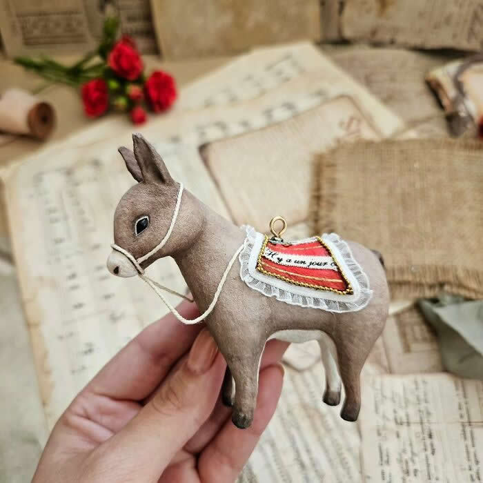 Miniature World: 33 Cotton Wool Sculptures by Katya Celebrating the Magic of Handmade Craft 59 A handcrafted miniature donkey ornament made from cotton wool stands in profile with a delicate bridle and a decorative saddle blanket trimmed with lace, photographed against a vintage backdrop of sheet music, old letters, and red roses.