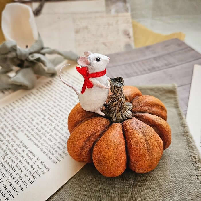 Miniature World: 33 Cotton Wool Sculptures by Katya Celebrating the Magic of Handmade Craft 60 A handcrafted miniature mouse ornament made from cotton wool stands on a rustic pumpkin, wearing a bright red scarf and posed beside vintage book pages and soft autumn décor.