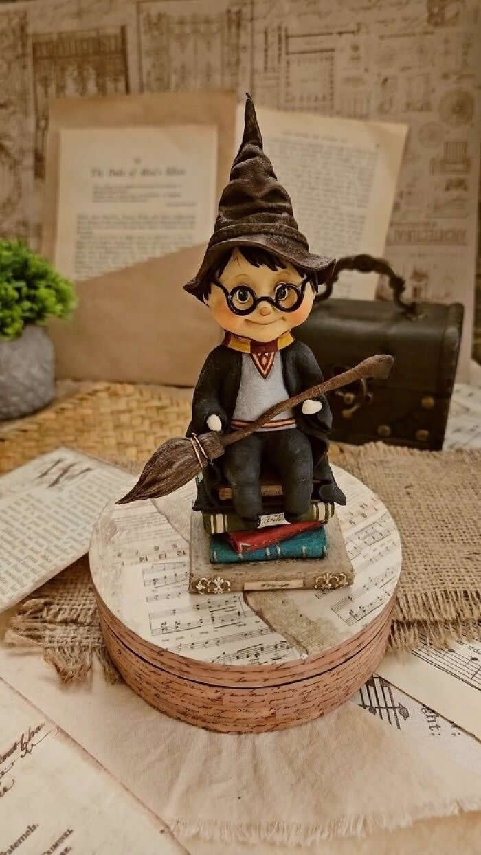 Miniature World: 33 Cotton Wool Sculptures by Katya Celebrating the Magic of Handmade Craft 61 A handcrafted miniature wizard boy ornament sits on a stack of tiny books, holding a broomstick and wearing round glasses and a pointed hat, displayed on a vintage-style tabletop with sheet music, old papers, and a small suitcase in the background.