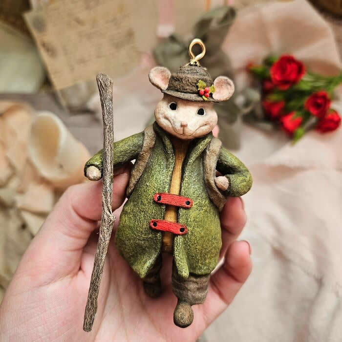 Miniature World: 33 Cotton Wool Sculptures by Katya Celebrating the Magic of Handmade Craft 62 A handcrafted miniature mouse ornament made from cotton wool is dressed in a moss-green coat, tiny brown hat, and rustic boots, holding a walking stick while standing in a hand against a soft vintage-style background with candles, fabric, and red roses.