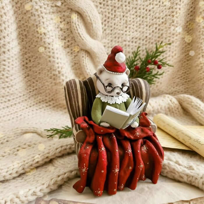 Miniature World: 33 Cotton Wool Sculptures by Katya Celebrating the Magic of Handmade Craft 63 A handcrafted cotton wool mouse ornament sits in a striped armchair reading a tiny book, dressed in a green outfit, lace collar, round glasses, and a red Santa hat, wrapped in a red patterned blanket against a cozy knitted winter backdrop.
