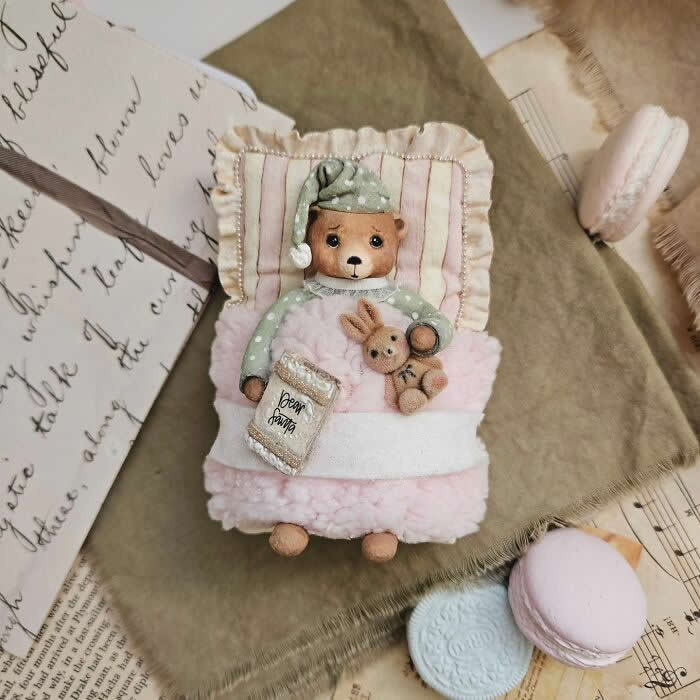 Miniature World: 33 Cotton Wool Sculptures by Katya Celebrating the Magic of Handmade Craft 64 A handcrafted cotton wool bear ornament lies tucked into a tiny pink bed, wearing mint-green polka-dot pajamas and a matching nightcap, cuddling a small bunny toy and holding a miniature bedtime book, surrounded by vintage papers and pastel macarons.