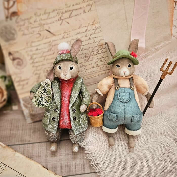 Miniature World: 33 Cotton Wool Sculptures by Katya Celebrating the Magic of Handmade Craft 65 Two handcrafted cotton wool rabbit ornaments stand side by side in a vintage-style scene—one dressed in a frosted winter coat holding a tiny tree, the other in blue overalls with a basket of red berries and a small pitchfork—set against soft fabric and antique paper backgrounds.