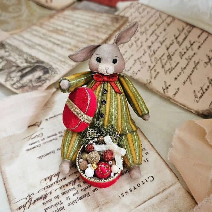 Miniature World: 33 Cotton Wool Sculptures by Katya Celebrating the Magic of Handmade Craft 67 A handcrafted cotton wool rabbit ornament in a striped green-and-gold outfit stands on vintage paper, holding a large red ornament and a tiny basket filled with miniature festive decorations, finished with a bright red bow at the neck.