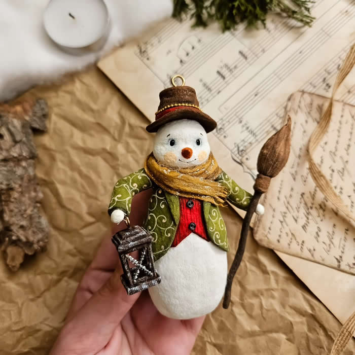 Miniature World: 33 Cotton Wool Sculptures by Katya Celebrating the Magic of Handmade Craft 41 Cotton Wool Sculptures. A handcrafted miniature snowman ornament made from cotton wool wears a green patterned coat, red vest, brown hat, and golden scarf while holding a lantern and a rustic broom against a vintage paper and sheet music backdrop.