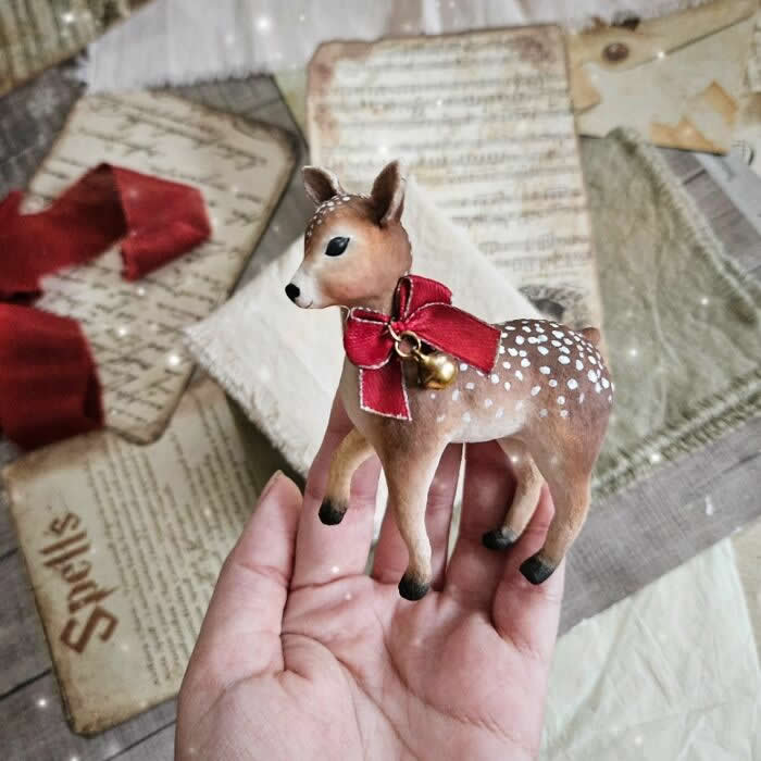 Miniature World: 33 Cotton Wool Sculptures by Katya Celebrating the Magic of Handmade Craft 68 A handcrafted cotton wool deer ornament with delicate white spots stands in a hand against a vintage paper background, wearing a large red ribbon with a tiny gold bell, evoking a soft and festive woodland holiday charm.