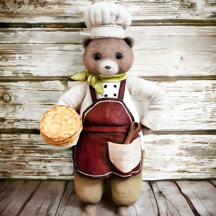 Miniature World: 33 Cotton Wool Sculptures by Katya Celebrating the Magic of Handmade Craft 69 A handcrafted cotton wool bear ornament dressed as a baker stands against a rustic wooden backdrop, wearing a white chef’s hat, green scarf, and burgundy apron while holding a stack of golden pastries, with a wooden spoon tucked into the apron pocket.