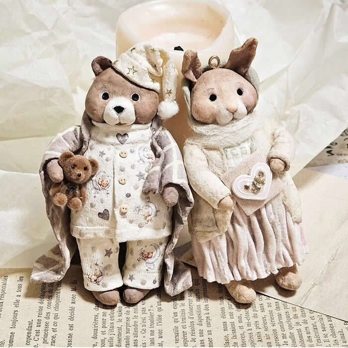 Miniature World: 33 Cotton Wool Sculptures by Katya Celebrating the Magic of Handmade Craft 70 Two handcrafted cotton wool ornaments stand side by side on vintage paper in a cozy holiday setting: a teddy bear in cream pajamas and a nightcap holding a tiny teddy, and a rabbit in a soft white sweater and pleated skirt holding a heart-shaped keepsake.