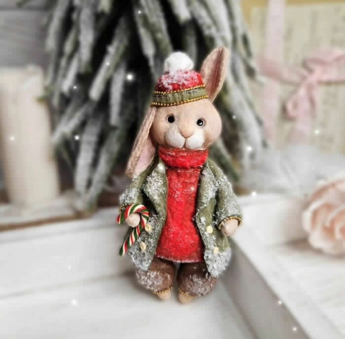 Miniature World: 33 Cotton Wool Sculptures by Katya Celebrating the Magic of Handmade Craft 71 A handcrafted cotton wool rabbit ornament stands in a snowy holiday scene, dressed in a frosted green coat, bright red scarf, and knit winter cap, holding a tiny candy cane beside a blurred Christmas tree and soft candlelit background.