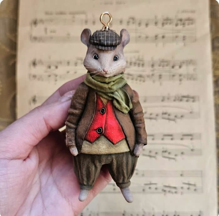 Miniature World: 33 Cotton Wool Sculptures by Katya Celebrating the Magic of Handmade Craft 44 A handcrafted miniature mouse ornament made from cotton wool wears a brown coat, red waistcoat, olive scarf, and plaid cap, standing against a softly blurred sheet music background with a charming old-world feel.