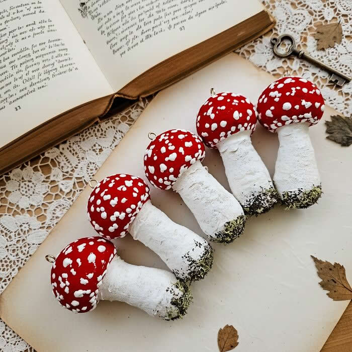 Miniature World: 33 Cotton Wool Sculptures by Katya Celebrating the Magic of Handmade Craft 45 Five handcrafted fly agaric mushroom ornaments made from cotton wool are arranged in a row on a lace-covered surface, with bright red caps, white speckles, and textured stems styled beside an open vintage book.