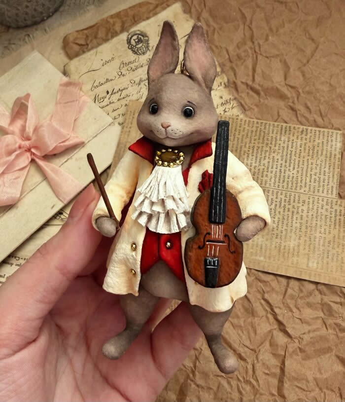 Miniature World: 33 Cotton Wool Sculptures by Katya Celebrating the Magic of Handmade Craft 47 A handcrafted miniature rabbit ornament made from cotton wool wears an elegant cream coat with red accents and a ruffled cravat, holding a tiny violin and bow against a vintage paper background.
