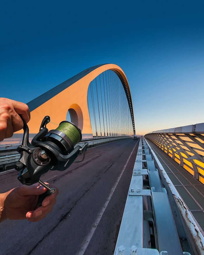 Pure Visual Magic: 35 Mind-Bending Forced Perspective Photos by Hugo Suíssas 48 A hand holds a fishing reel in the foreground, aligned with a large arched bridge at sunset so the bridge appears to be threaded through the reel like a fishing line in a playful forced perspective illusion.