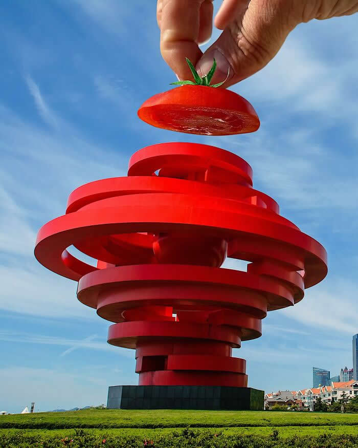 Pure Visual Magic: 35 Mind-Bending Forced Perspective Photos by Hugo Suíssas 49 A hand holds a sliced tomato above a large red spiral sculpture, aligned so the tomato appears to become the top layer of the structure in a bold forced perspective illusion against a blue sky.