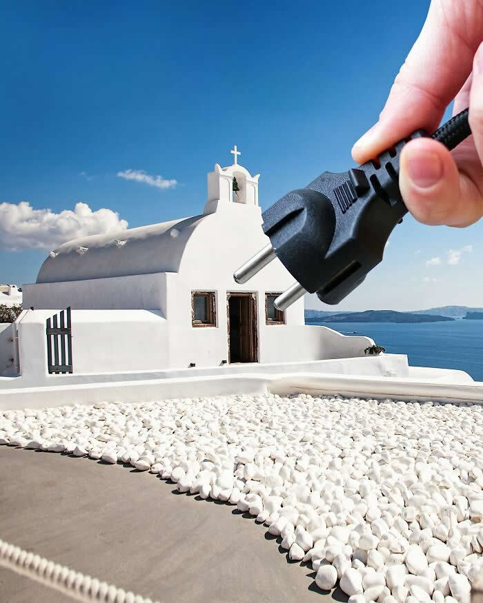 Pure Visual Magic: 35 Mind-Bending Forced Perspective Photos by Hugo Suíssas 50 A hand holds a black electrical plug in the foreground, aligned with a white seaside chapel so the plug appears to connect directly into the building, creating a playful forced perspective illusion against the blue sky and sea.