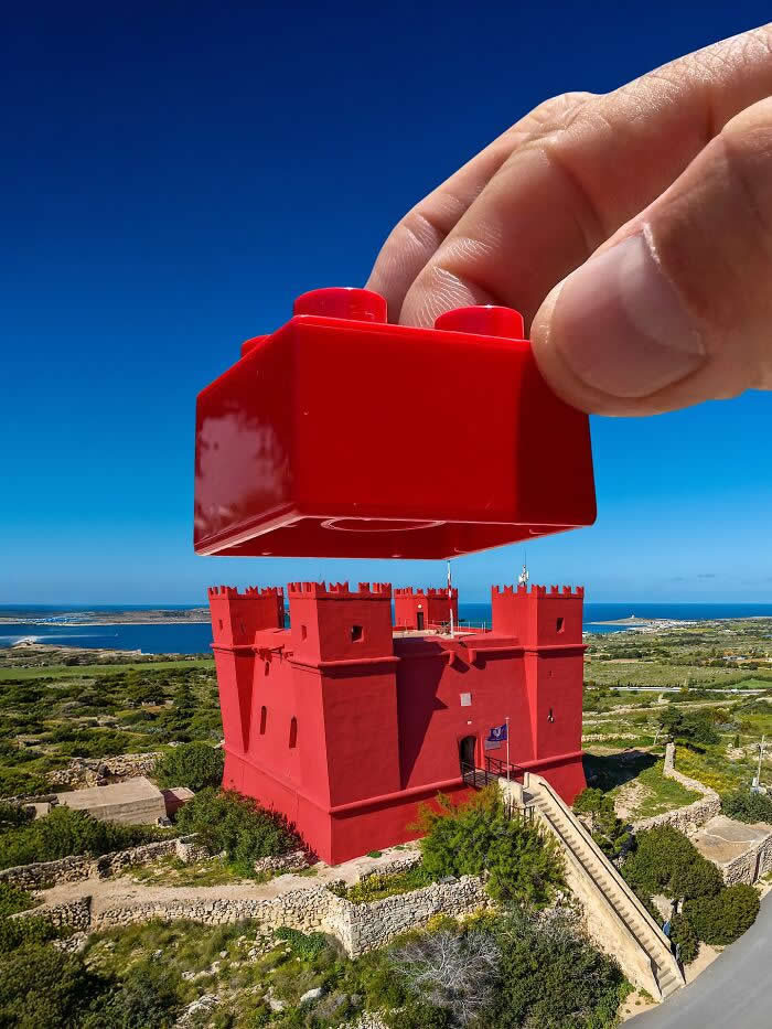 Pure Visual Magic: 35 Mind-Bending Forced Perspective Photos by Hugo Suíssas 56 A hand holds a red toy building block above a bright red fortress, aligned so the block appears to become the castle’s missing top piece in a playful forced perspective illusion against a vivid blue sky.