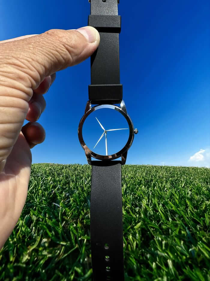 Pure Visual Magic: 35 Mind-Bending Forced Perspective Photos by Hugo Suíssas 40 A hand holds a black wristwatch against a bright blue sky and grassy field, aligning the watch face with a distant wind turbine so the turbine blades appear to become the hands of the watch in a playful forced perspective illusion.