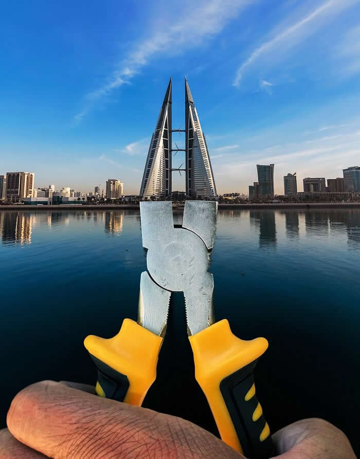 Pure Visual Magic: 35 Mind-Bending Forced Perspective Photos by Hugo Suíssas 63 A hand holds a pair of yellow-handled pliers in the foreground, aligned with the twin sail-shaped Bahrain World Trade Center towers so the pliers appear to grip the skyscrapers in a playful forced perspective illusion above calm waterfront reflections.