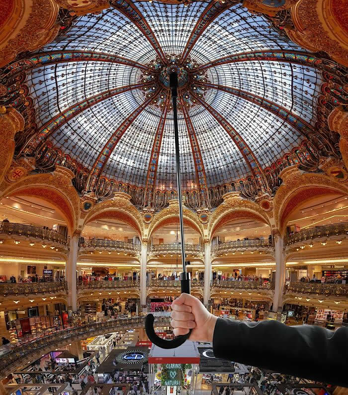 Pure Visual Magic: 35 Mind-Bending Forced Perspective Photos by Hugo Suíssas 65 Inside an ornate shopping gallery with a stained-glass dome, a hand holds an umbrella upward, perfectly aligned so the umbrella appears to support the entire ceiling in a playful forced perspective illusion.
