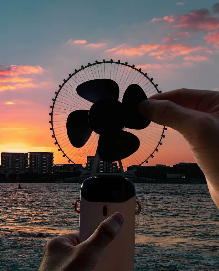 Pure Visual Magic: 35 Mind-Bending Forced Perspective Photos by Hugo Suíssas 66 At sunset by the water, a hand holds a small portable fan aligned with a giant Ferris wheel in the background, making the wheel look like the fan’s oversized spinning blades in a clever forced perspective composition.