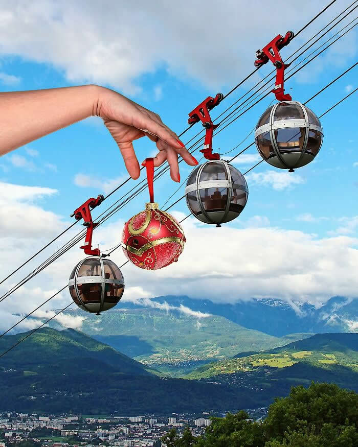 Pure Visual Magic: 35 Mind-Bending Forced Perspective Photos by Hugo Suíssas 68 A hand holds a red Christmas ornament in front of a cable car line, aligning it between the gondolas so it looks like the ornament is hanging from the same cable among the cabins in a clever forced perspective scene above a mountain landscape.