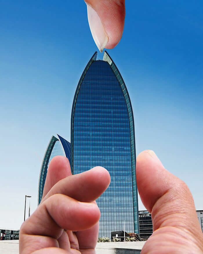 Pure Visual Magic: 35 Mind-Bending Forced Perspective Photos by Hugo Suíssas 69 Two hands frame a tall curved glass skyscraper against a clear blue sky, aligned so the fingertips appear to pinch the pointed top of the building in a playful forced perspective illusion.