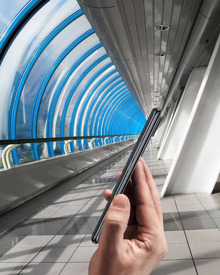 Pure Visual Magic: 35 Mind-Bending Forced Perspective Photos by Hugo Suíssas 44 A hand holds a slim smartphone inside a curved glass walkway, aligned so the phone appears to fold shut around the tunnel, turning the arched passage into part of a playful forced perspective illusion.