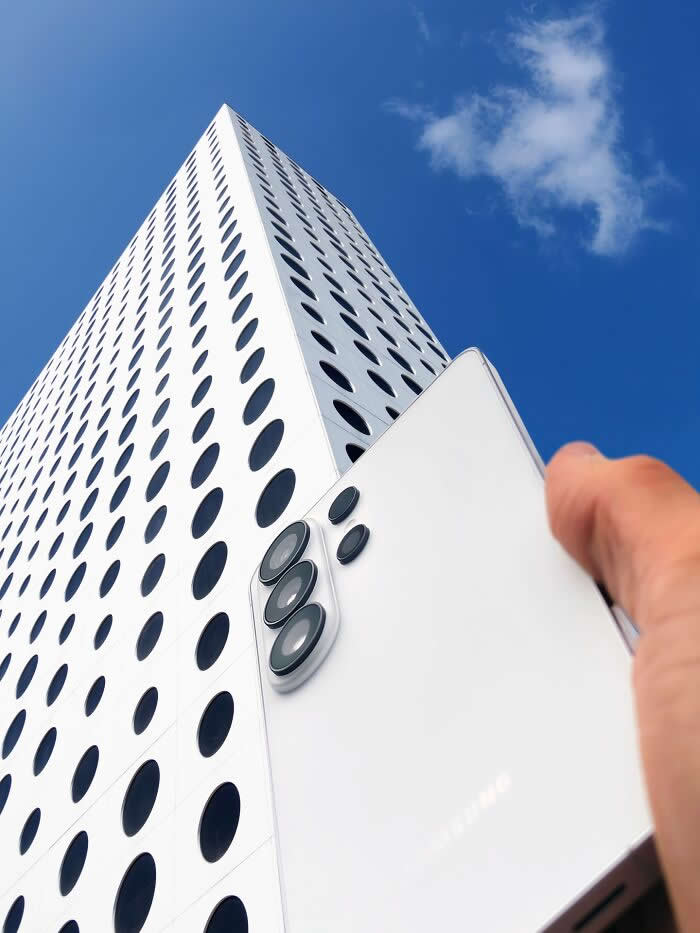 Pure Visual Magic: 35 Mind-Bending Forced Perspective Photos by Hugo Suíssas 45 A hand holds a white smartphone in front of a tall white building with circular black windows, aligning the phone’s rear camera lenses with the building’s dotted façade to create a playful forced perspective illusion.