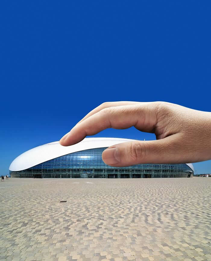 Pure Visual Magic: 35 Mind-Bending Forced Perspective Photos by Hugo Suíssas 46 A hand hovers above a sleek oval-shaped modern building, aligned so the fingers appear to gently pinch or press the curved white roof against a deep blue sky in a clever forced perspective composition.