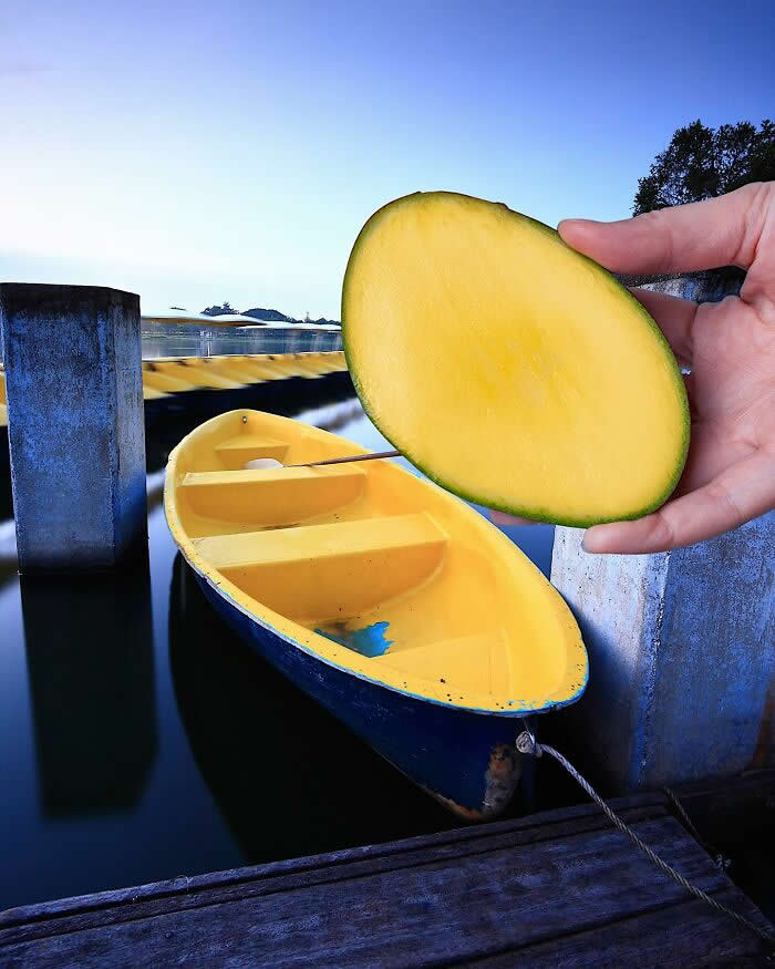 Pure Visual Magic: 35 Mind-Bending Forced Perspective Photos by Hugo Suíssas 47 A hand holds half a mango in the foreground, perfectly aligned with a yellow boat floating in calm water so the fruit appears to become the boat’s upper half in a playful forced perspective illusion.